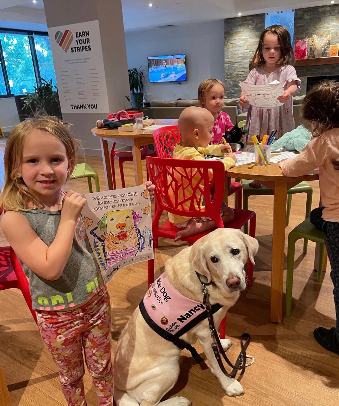 Child holding up a picture of a dog she coloured in, while standing next to Nancy the Retired Guide Dog as part of Learning Centre activities at Ronald McDonald House Greater Western Sydney Westmead